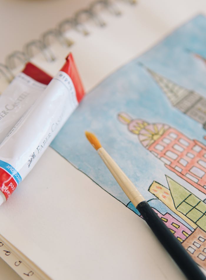 Notebook open to a watercolor painting of cityscape with paintbrush and tube in view.