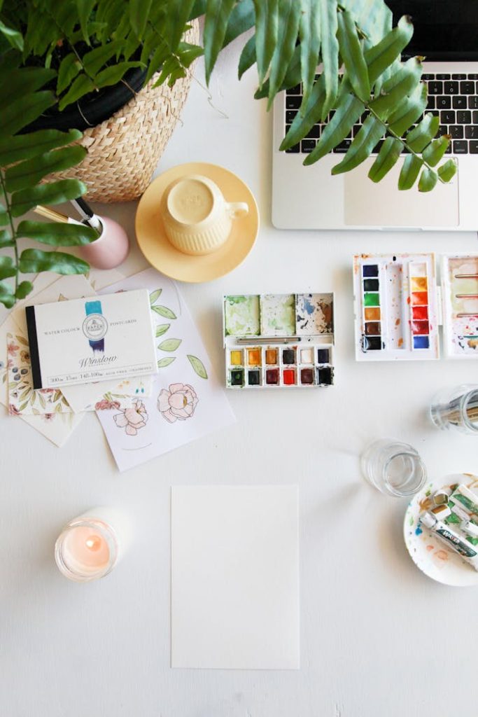 Flat lay of a creative workspace with watercolors, plants, and a laptop.
